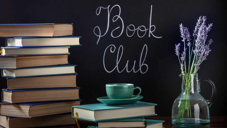 Stack of books with coffee mg on top, lilacs in vase and Book Club written on a chalk board
